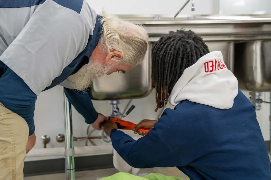 Two people work together under a metal sink, using a wrench to adjust plumbing pipes—honing skills that are essential in plumbing technician courses.