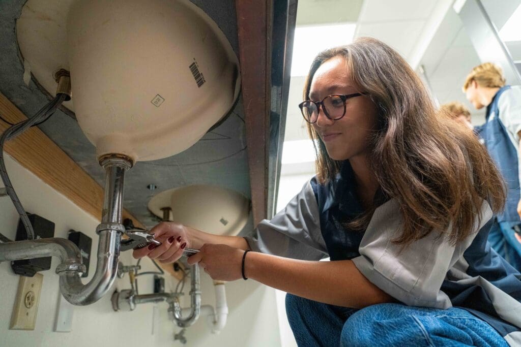 A person uses a wrench to adjust pipes under a sink in a classroom or workshop setting.