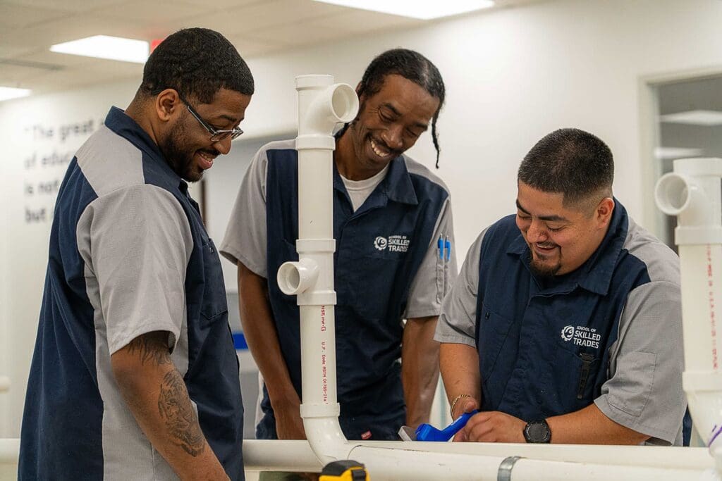 Three men in matching uniforms work together on assembling PVC pipes indoors, appearing focused and collaborative.