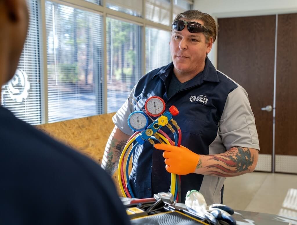 A technician wearing orange gloves and safety glasses holds a manifold gauge set in a vocational school workshop, speaking to another person.